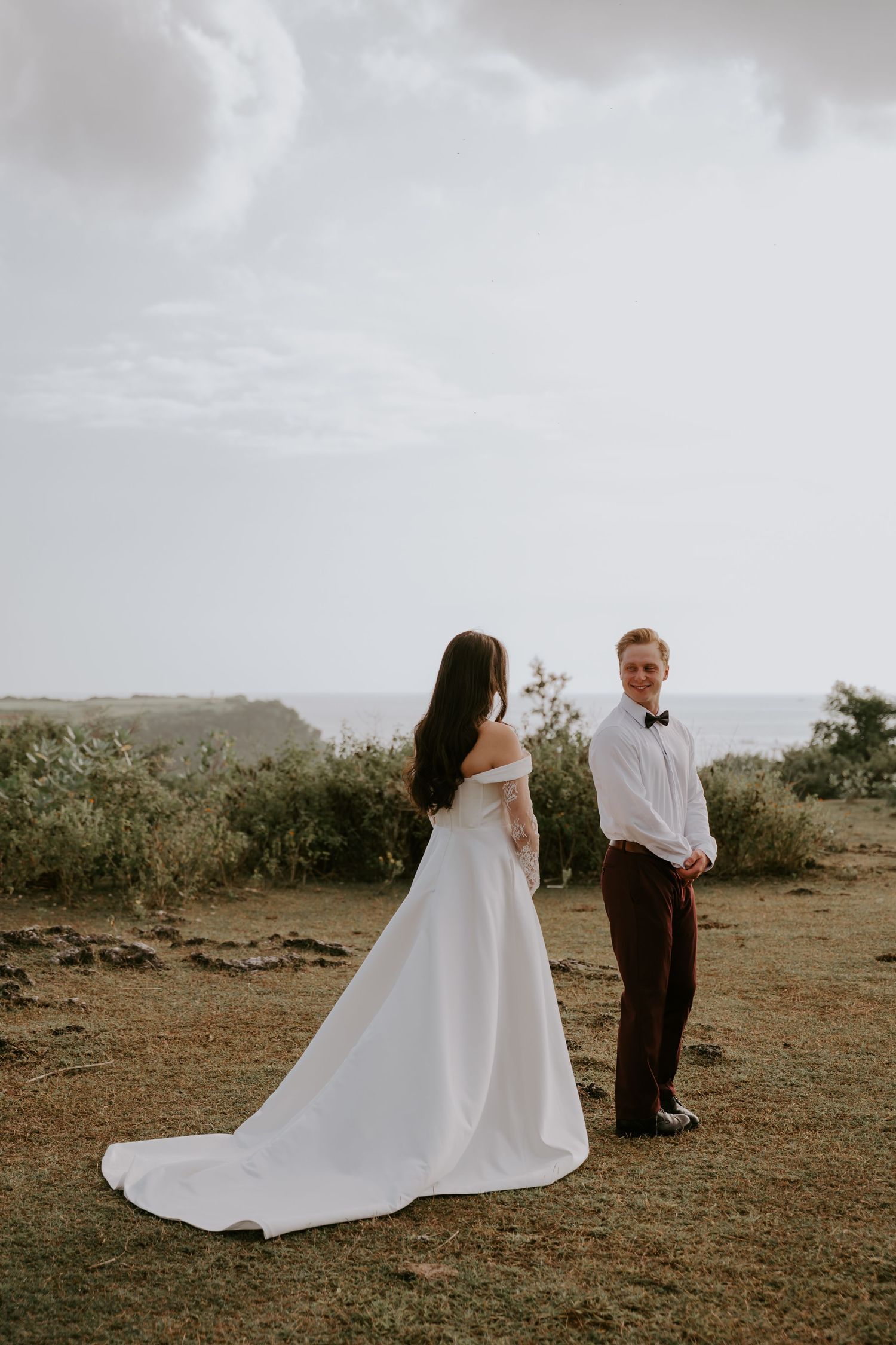 Bride in off-shoulder white gown faces groom in white shirt and bow tie during outdoor wedding ceremony by the ocean.