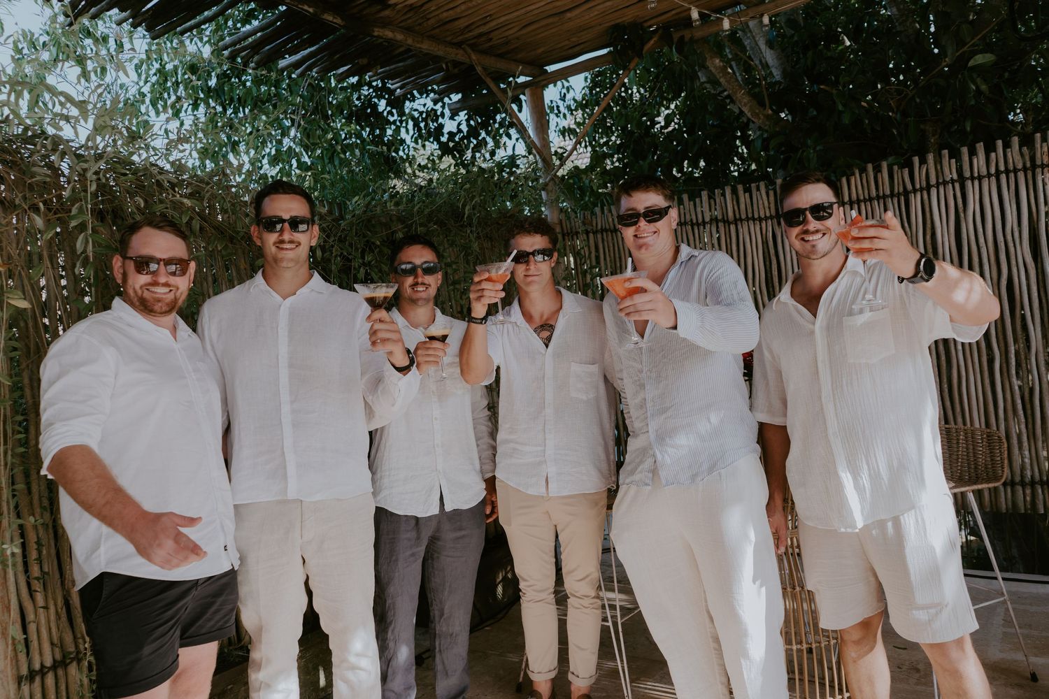 Six groomsmen in white linen shirts raising glasses in celebratory toast at outdoor wedding venue.