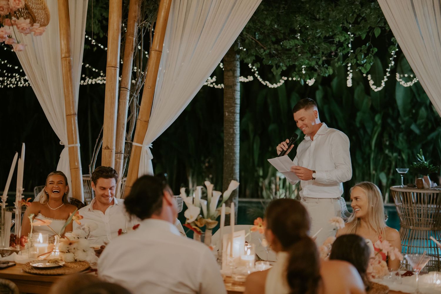 Wedding guest delivers speech to candlelit reception under bamboo pergola with string light decor.