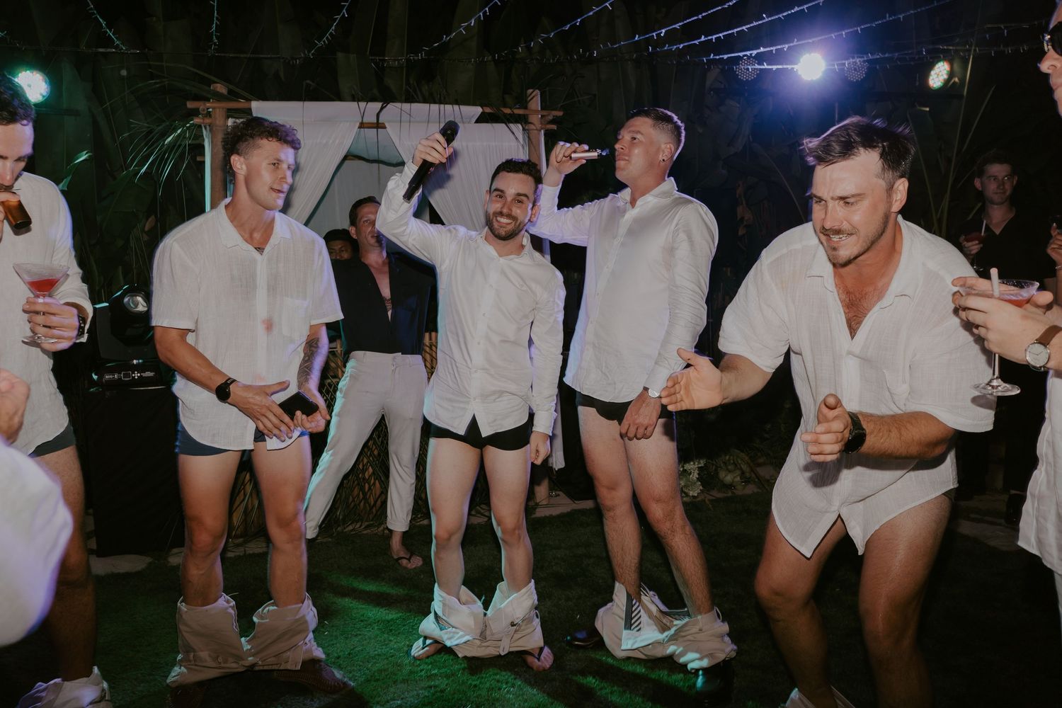 Group of male wedding guests in white shirts celebrate on the dance floor at an outdoor reception.