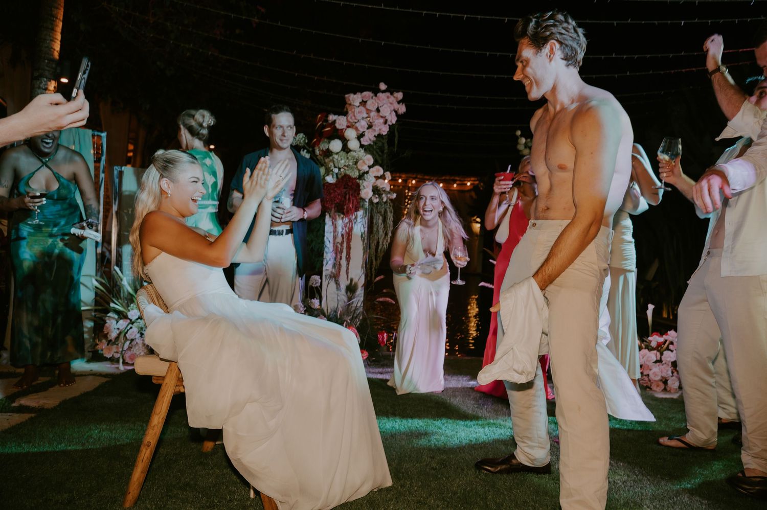 Shirtless groom performing garter removal while bride sits in chair at outdoor wedding reception.
