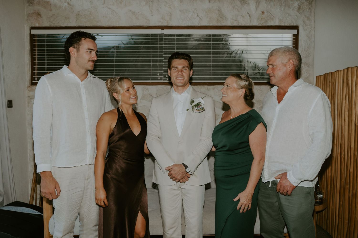Groom in white suit shares a joyful moment with family members inside a warmly lit rustic wedding venue.