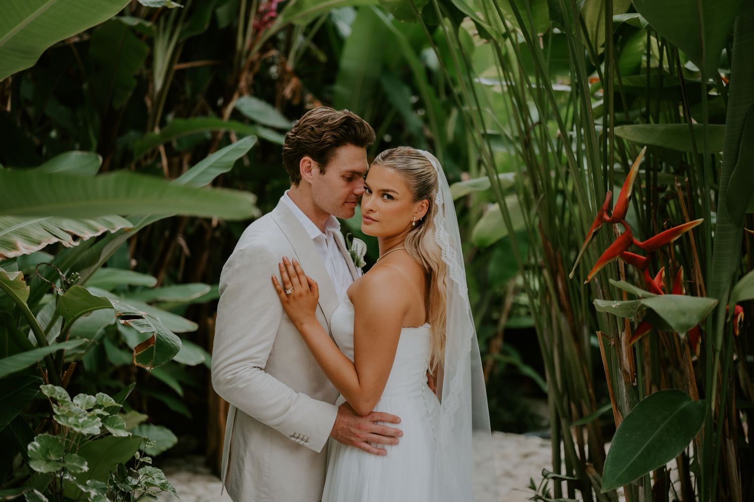 Newlywed couple embraces romantically among lush tropical banana leaves and bird of paradise plants.