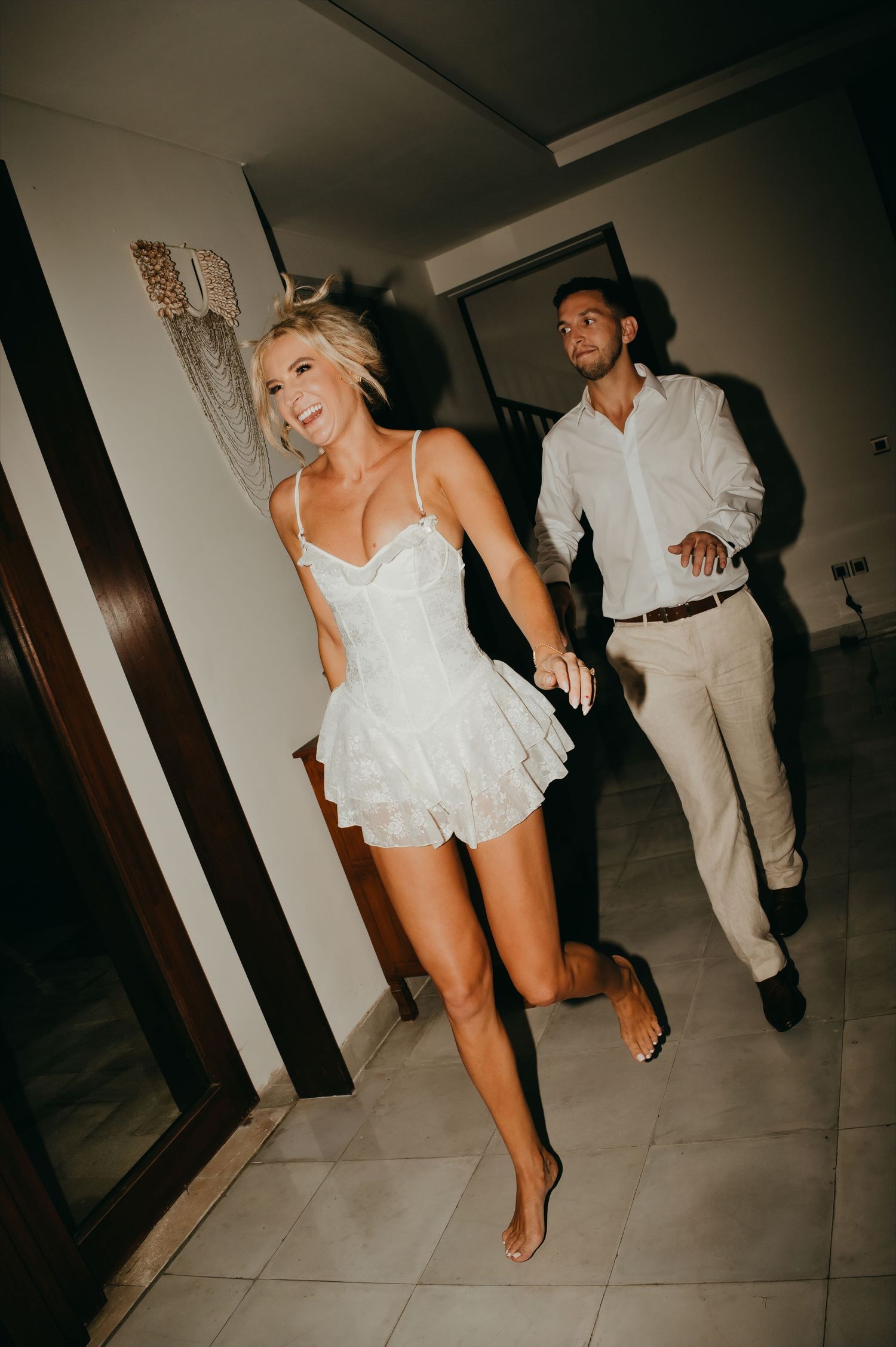 Bride in a white lace mini dress runs barefoot through a hallway with groom following close behind.