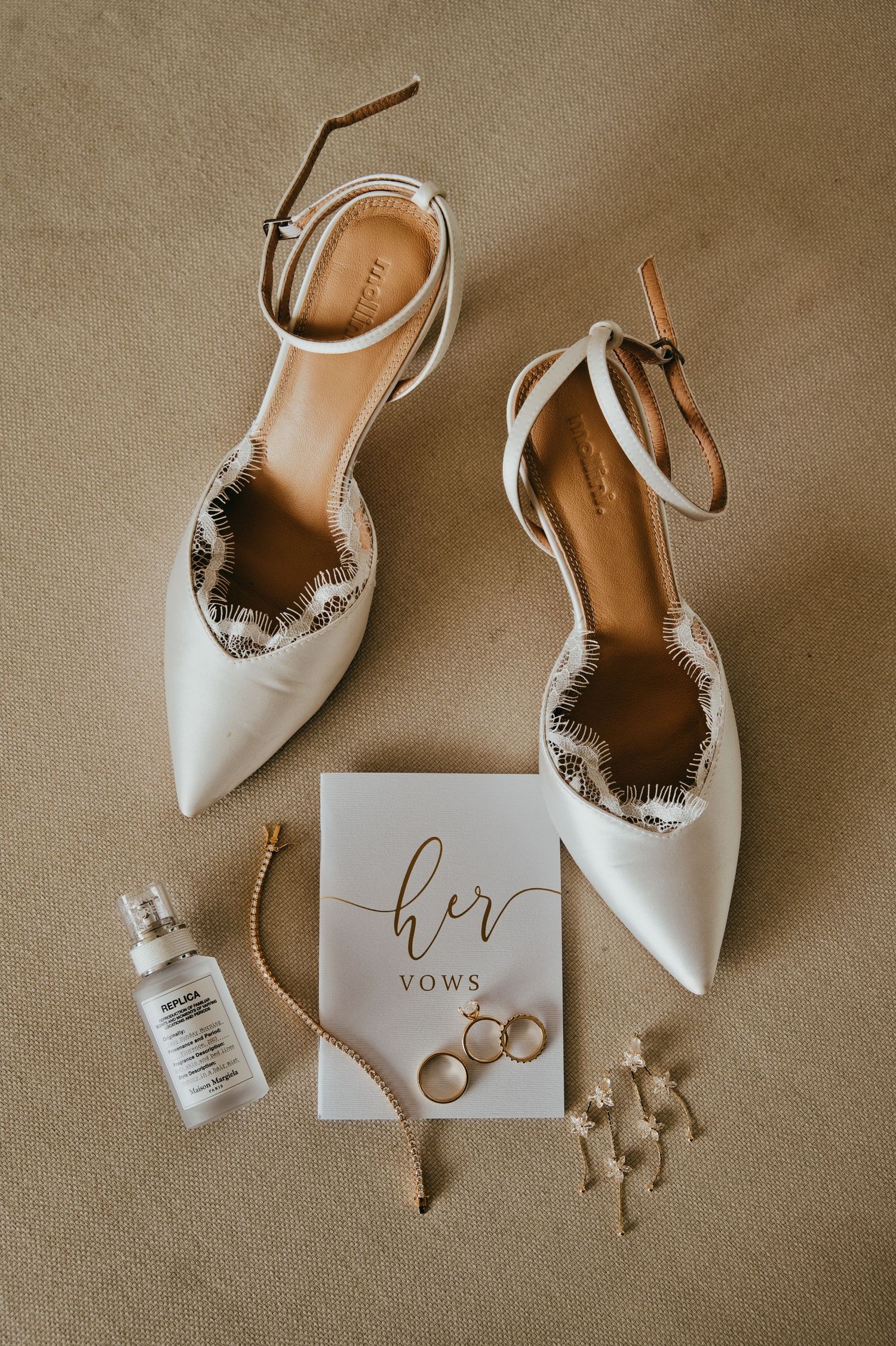 Bridal flat lay with white heels, vow book, wedding rings, earrings, and perfume.