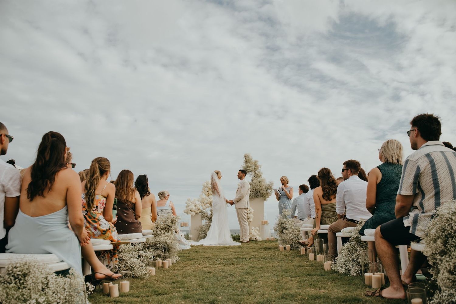 Bride and groom exchange vows at an outdoor clifftop ceremony with white floral arch and seated guests.