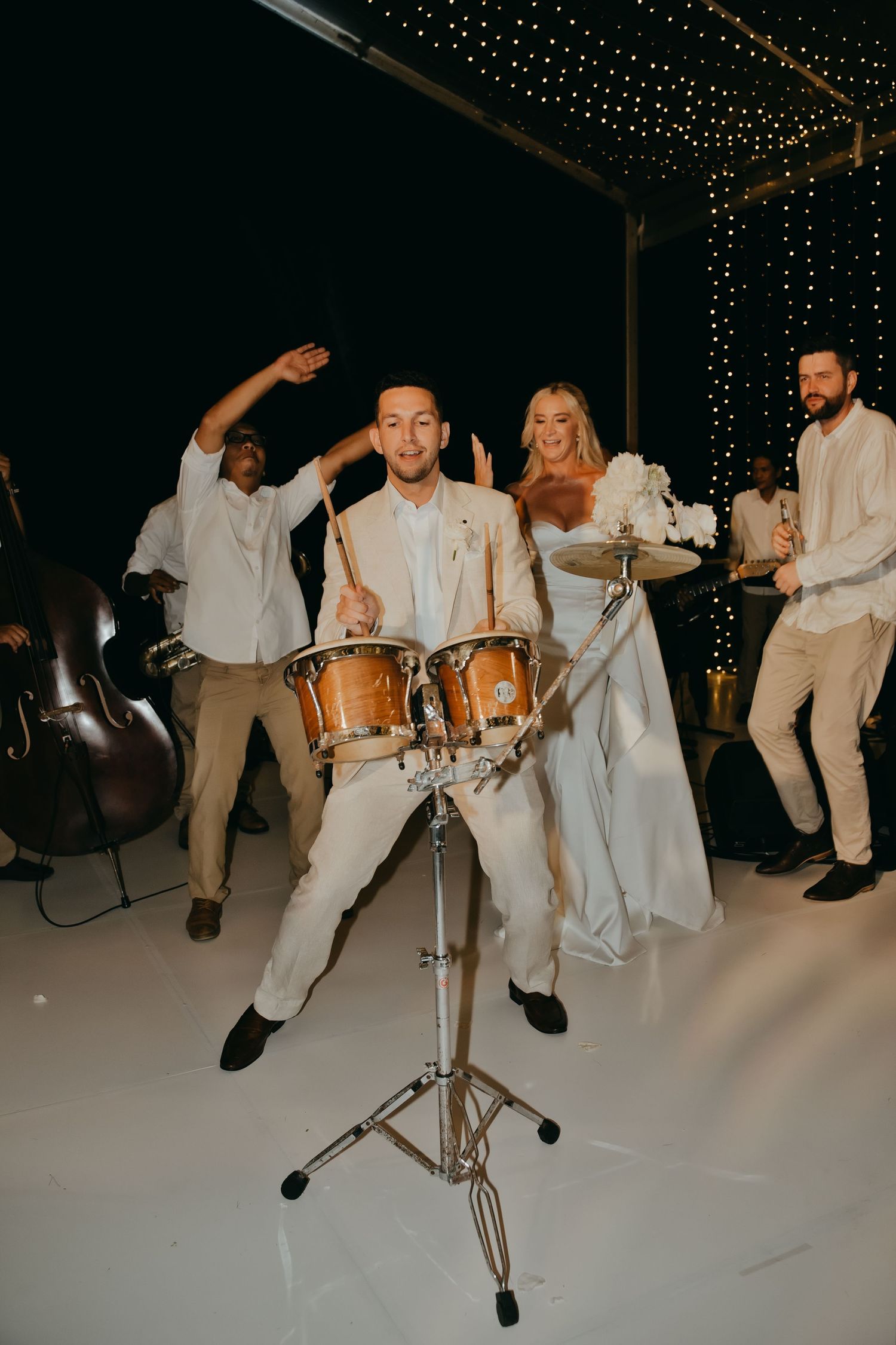 Groom plays bongo drums beside bride in white gown at lively wedding reception under fairy lights.