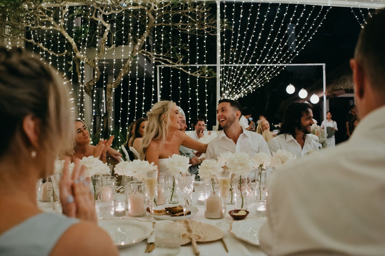 Bride and groom laugh together at elegant wedding reception table adorned with white floral centerpieces and lights.
