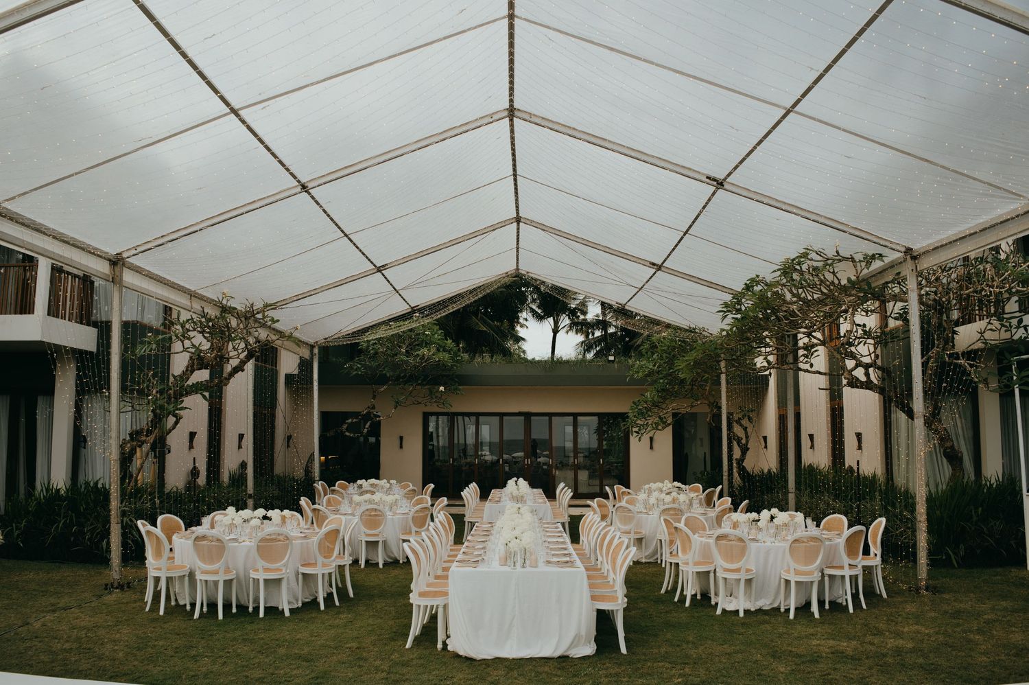 Elegant outdoor wedding reception under a clear marquee tent with round tables and white floral arrangements.