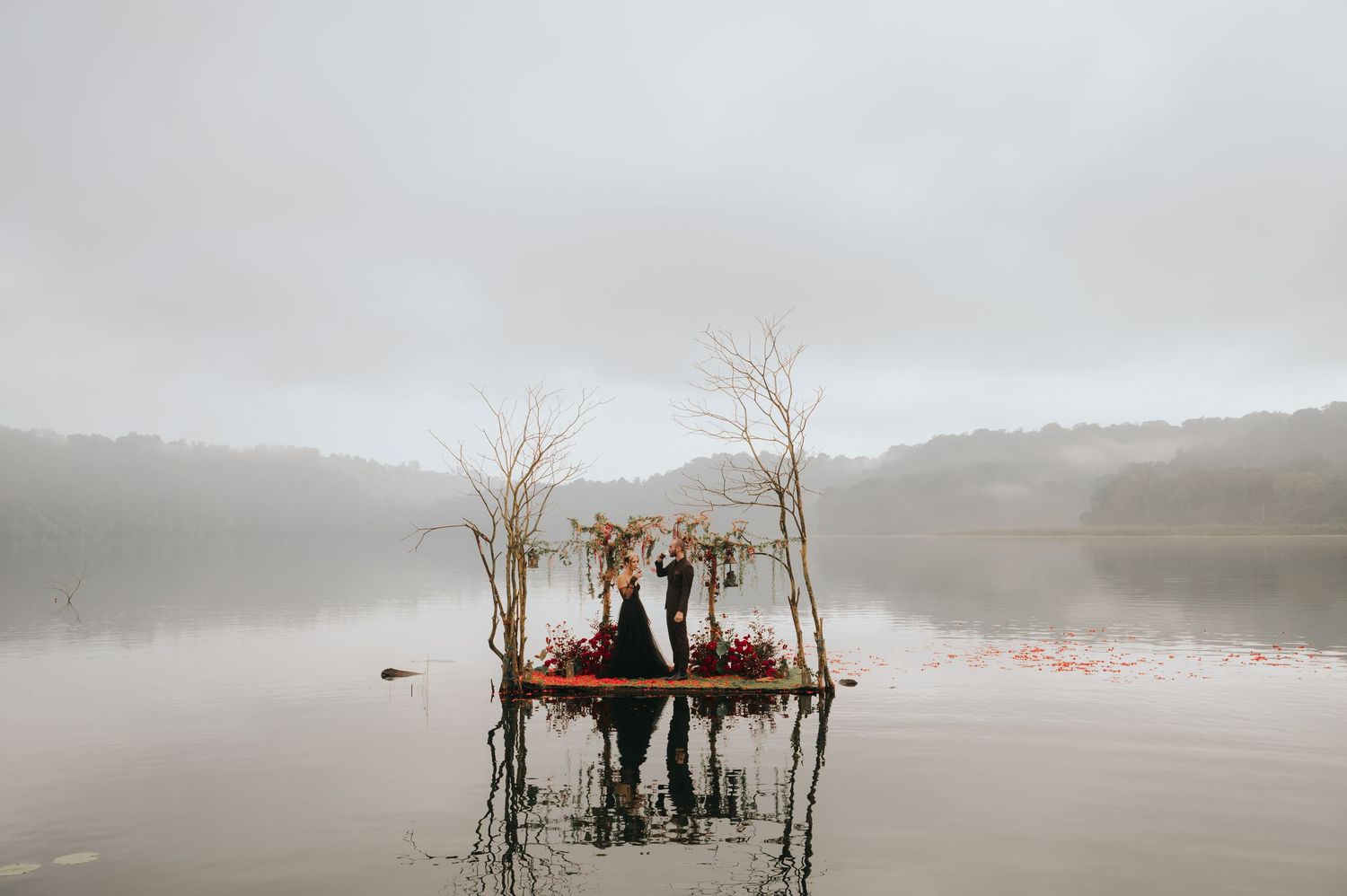 Silhouetted figures stand on a partially submerged platform in a foggy lake during golden hour.