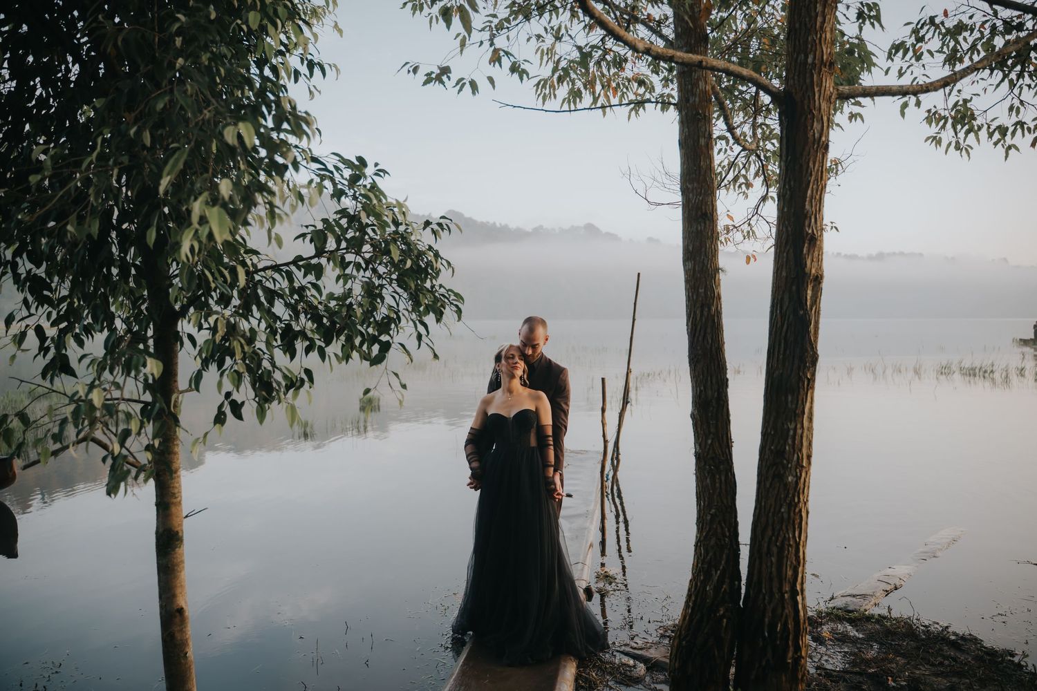 A romantic lakeside photoshoot with tall trees framing the misty water and mountains in the background.