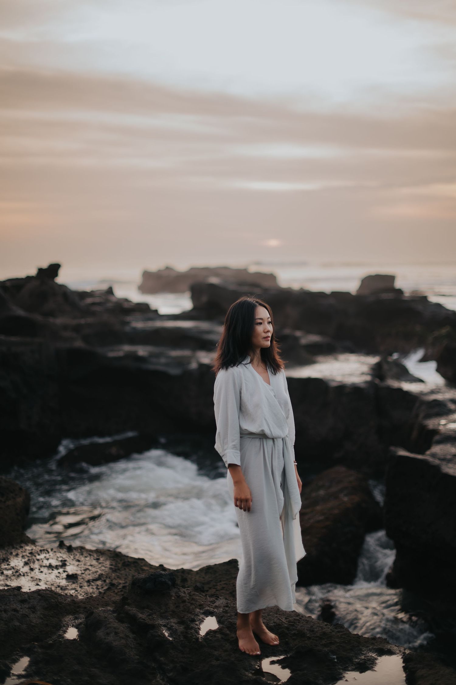 A figure in a flowing white dress stands on dark volcanic rocks at sunset along a dramatic coastal shoreline.
