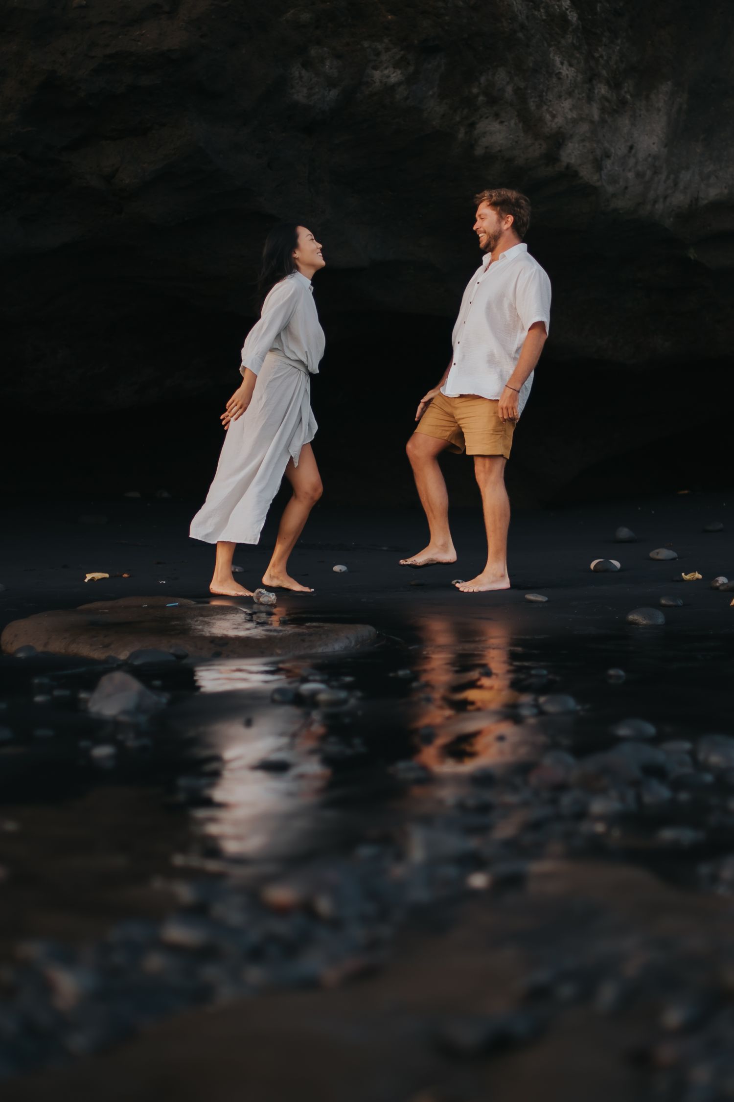 Two people wade in shallow water at dusk, creating reflections on the dark rocky shore.