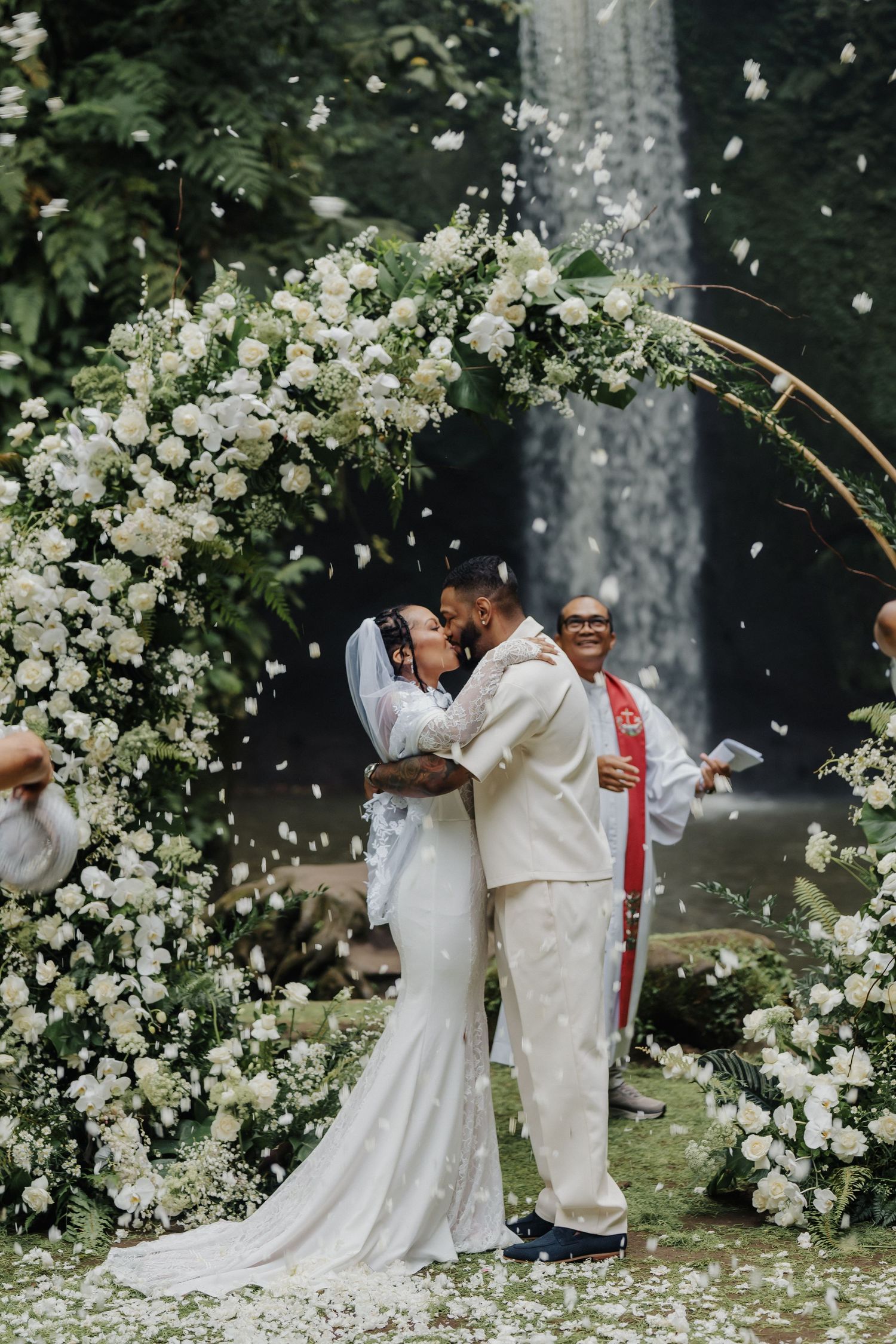 Bride and groom share first kiss under circular floral arch with white flowers and cascading waterfall background.