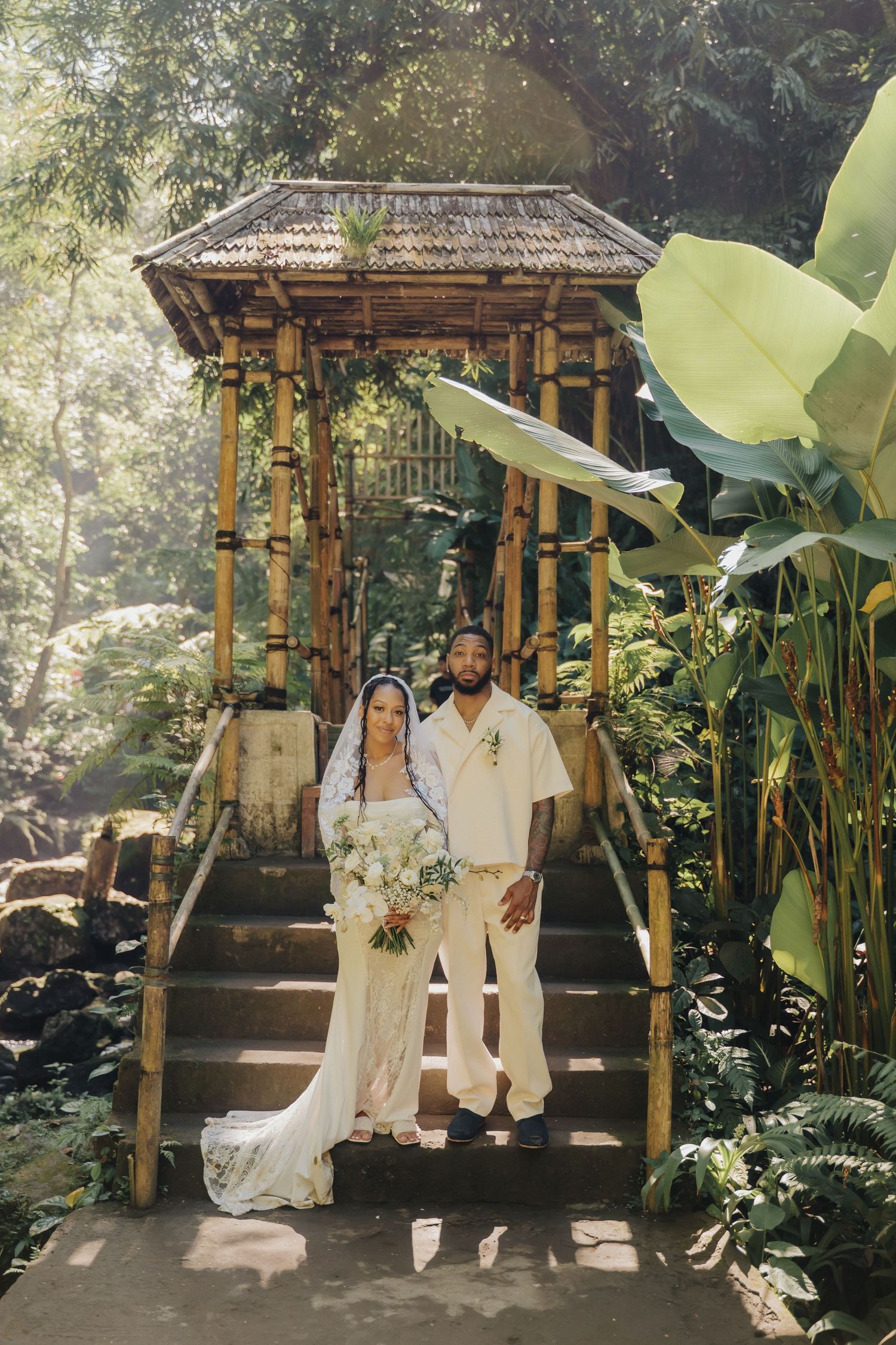 Wedding couple poses on stone steps before a bamboo gazebo surrounded by lush tropical greenery and banana leaves.