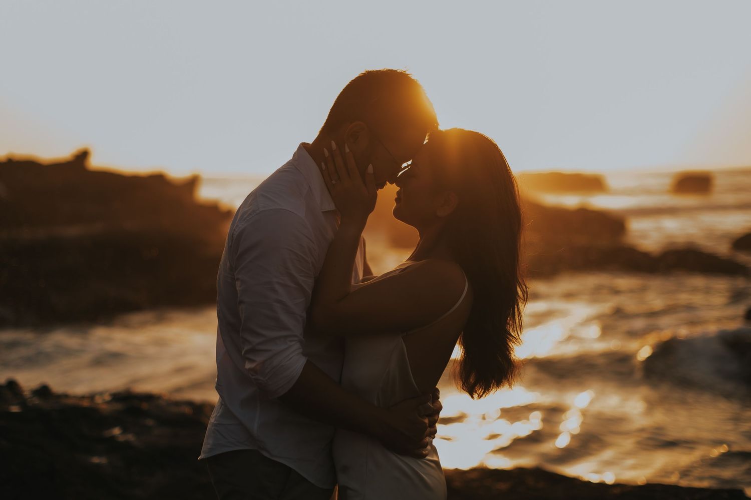 Couple sharing a romantic sunset kiss on rocky coastline with golden ocean light behind them.