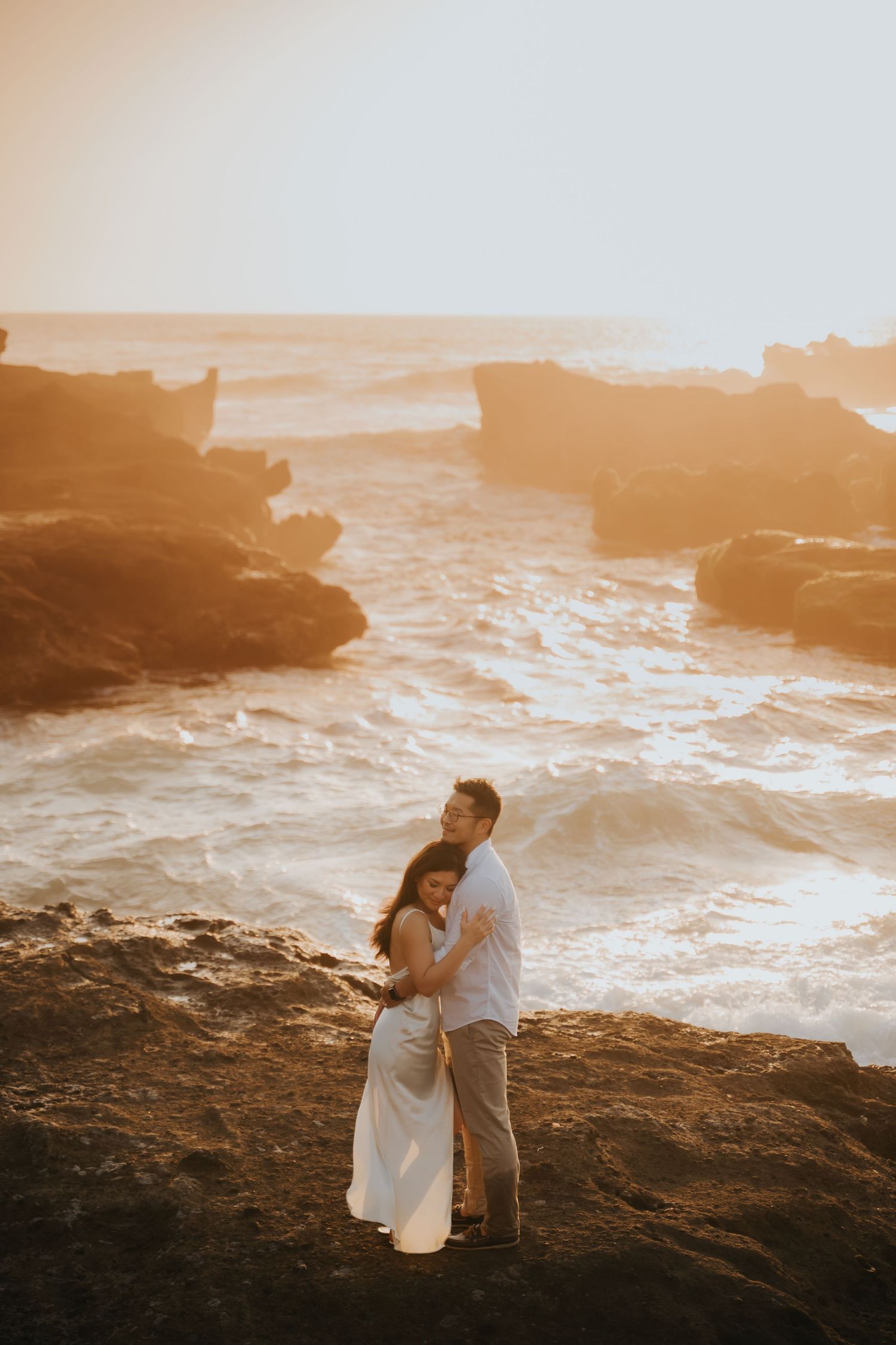 Couple embracing on rocky coastal cliffs at golden sunset with waves crashing behind them.