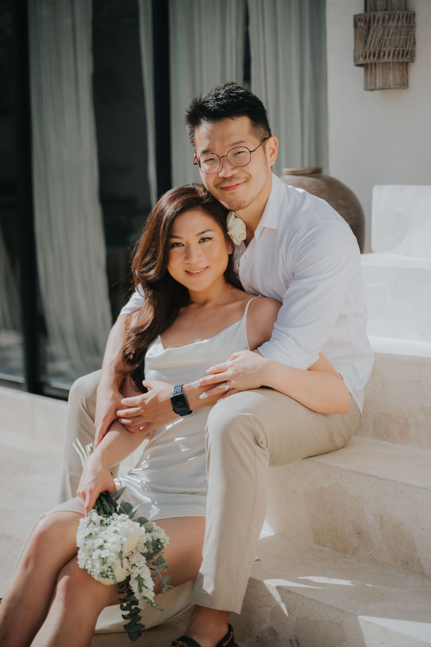 Couple in white outfits posing together, woman holding white flowers, man with glasses embracing her on outdoor steps.