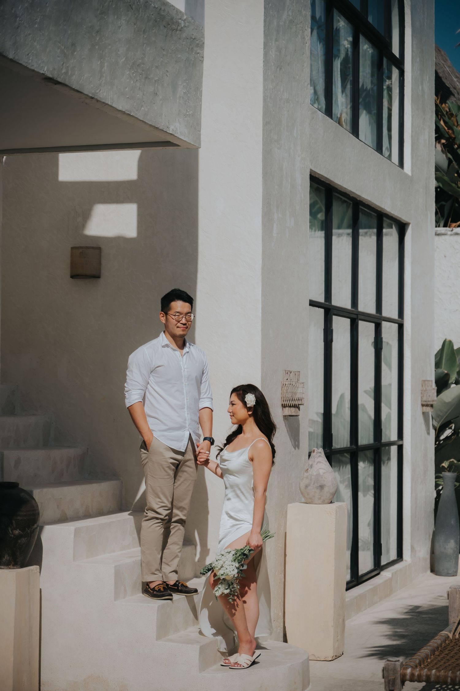 Couple in elegant attire holding hands outside a modern white villa, woman holding a bouquet with a flower in her hair.