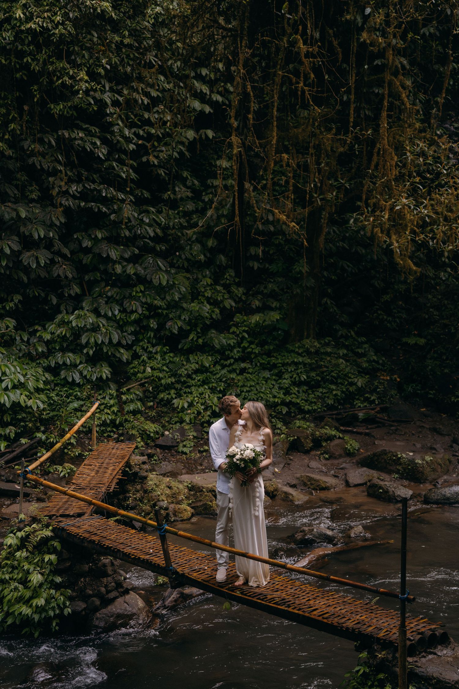 A couple stands on a rustic wooden bridge over a stream in a lush forest setting during a romantic moment.