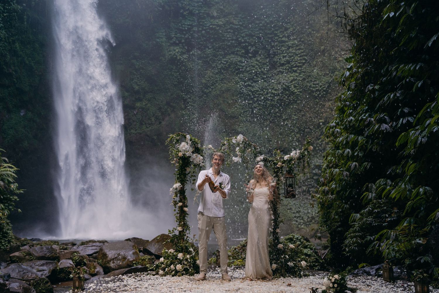 A couple stands together in front of a majestic waterfall in a lush forest setting.