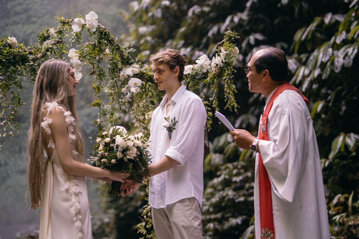 An outdoor wedding ceremony takes place under a floral arch with greenery and roses in a garden setting.