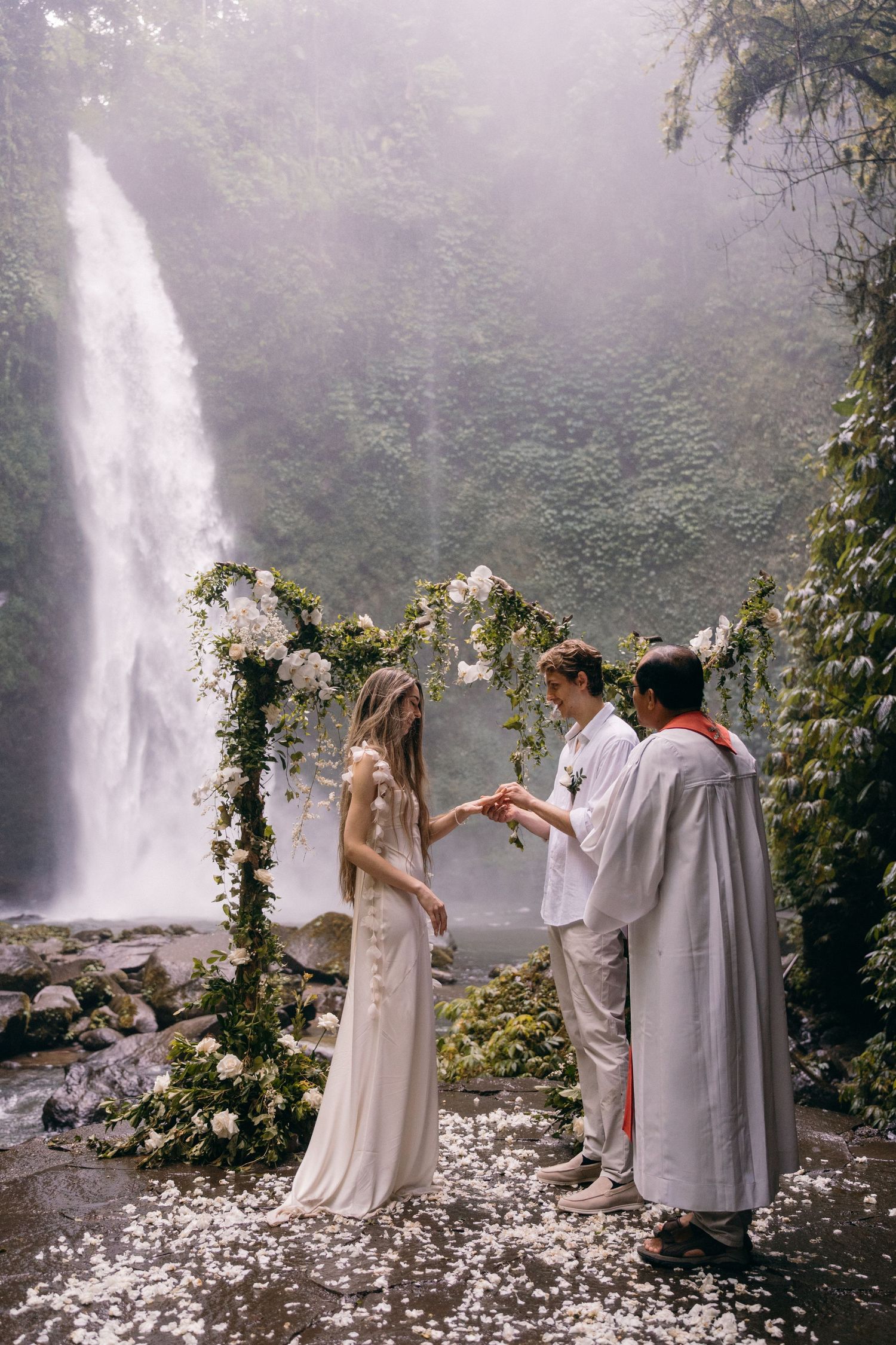A romantic ceremony takes place in front of a majestic waterfall with a floral arch and scattered petals on the ground.