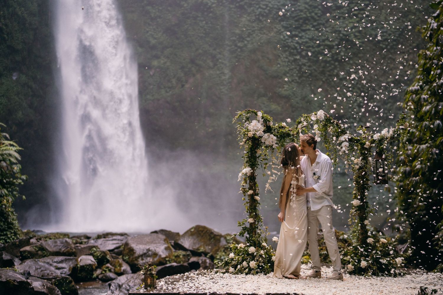 A couple shares an intimate moment under a floral arch with a majestic waterfall cascading in the background.