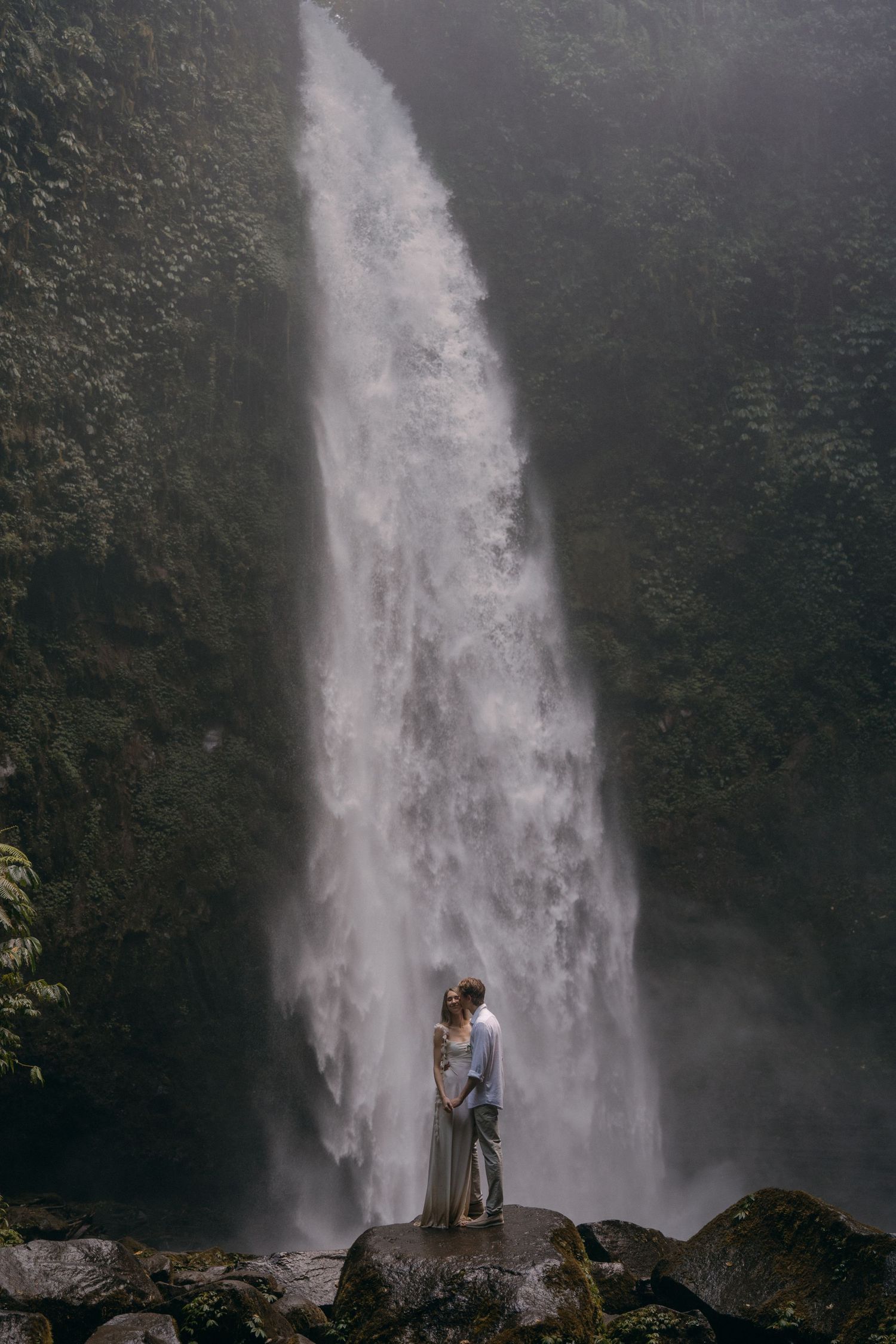 A couple stands in silhouette against the majestic backdrop of a towering waterfall cascading down a lush cliff face.