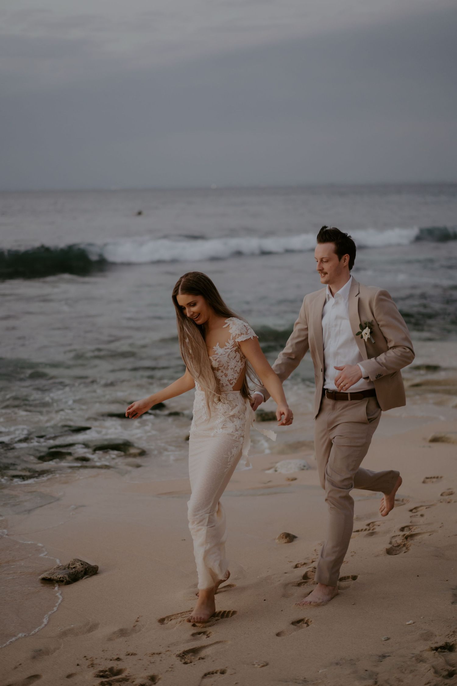 A couple in wedding attire playfully runs and dances together on a moody beach at sunset.