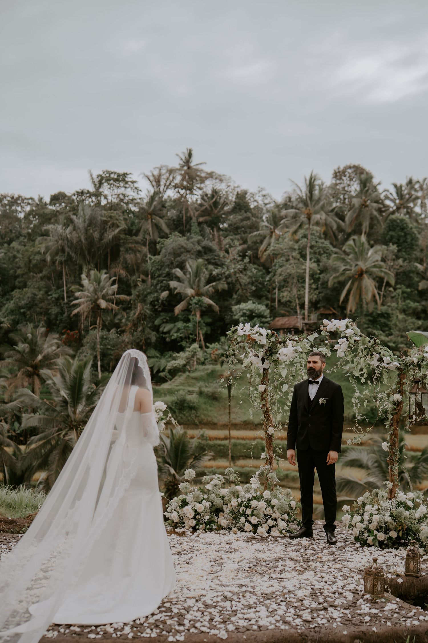 A wedding couple stands at an outdoor ceremony surrounded by palm trees and floral decorations in a tropical setting.