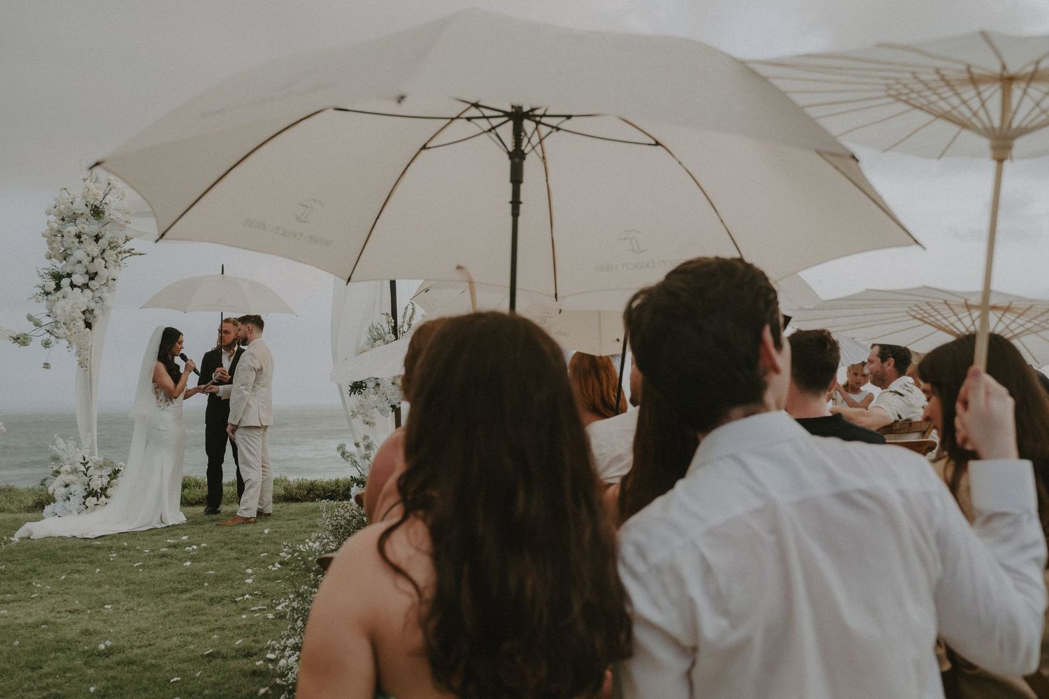 Wedding guests gather under white umbrellas during an outdoor ceremony on a misty hilltop.