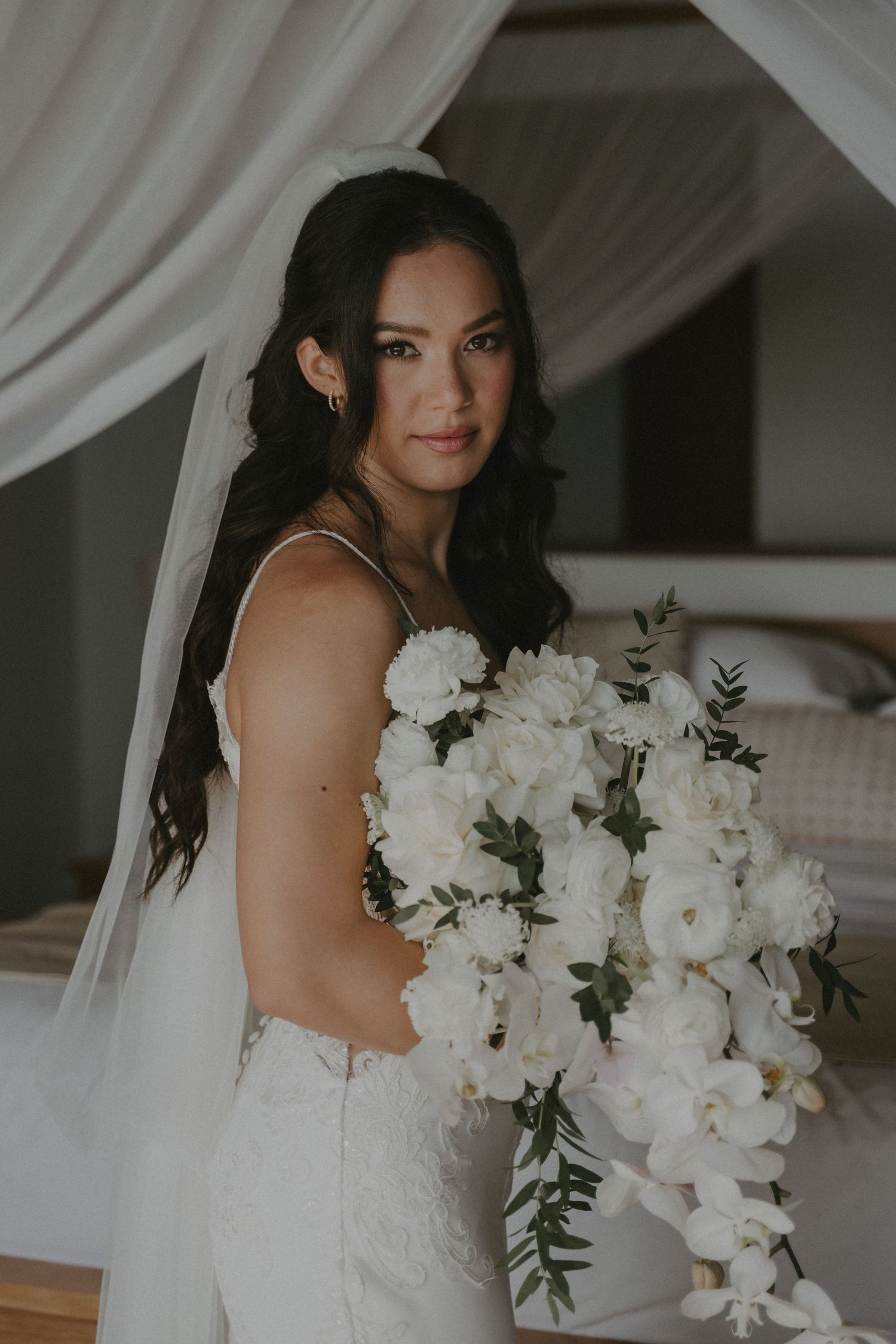 A bride in a white wedding dress holds an elegant bouquet of white flowers and eucalyptus against a light fabric backdrop.