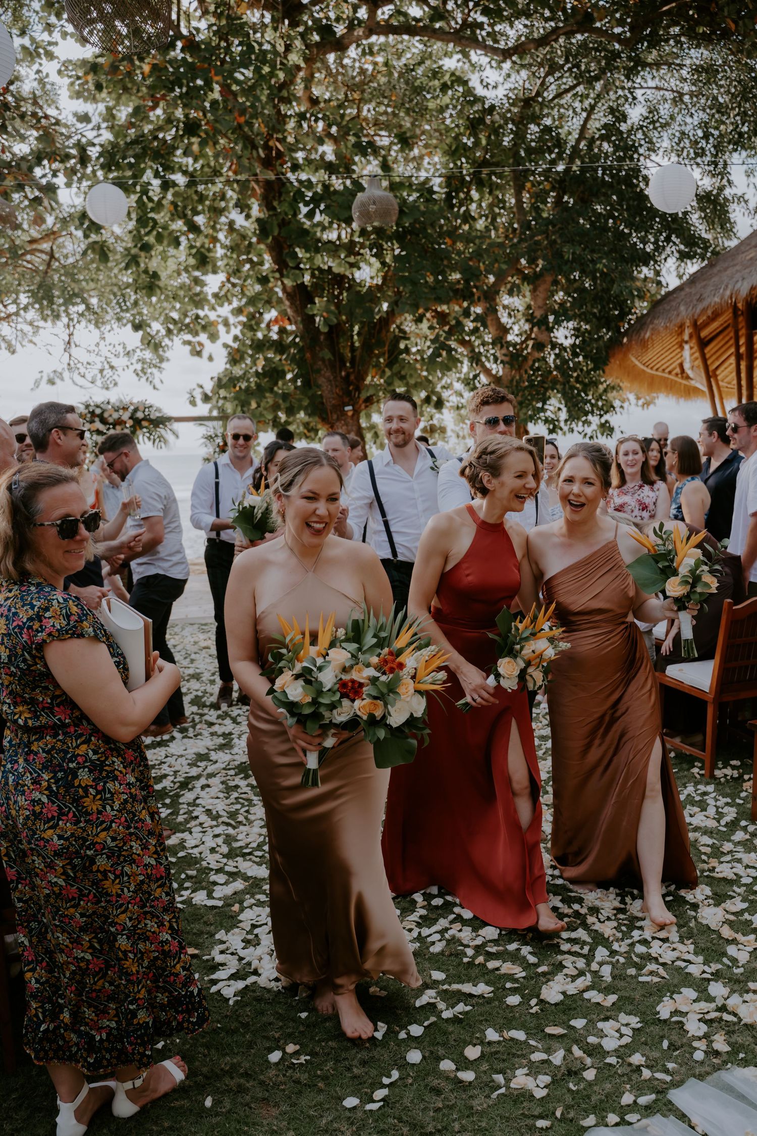 Wedding guests in colorful dresses gather on a lawn scattered with flower petals under a large tree at an outdoor celebration.