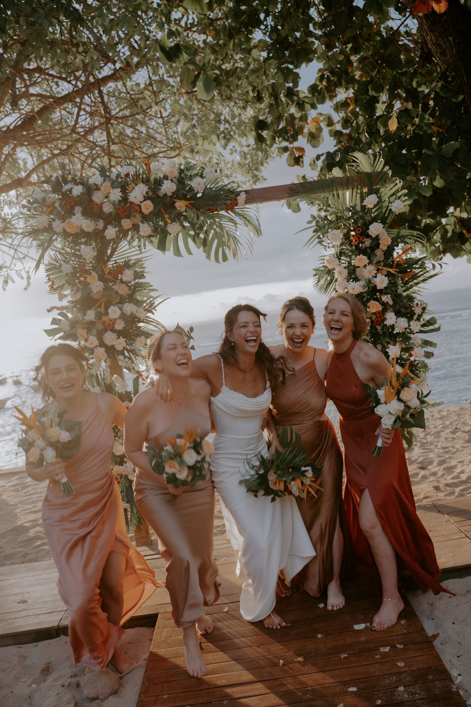 A group of bridesmaids in burgundy and blush dresses laughing together on a beach at sunset.