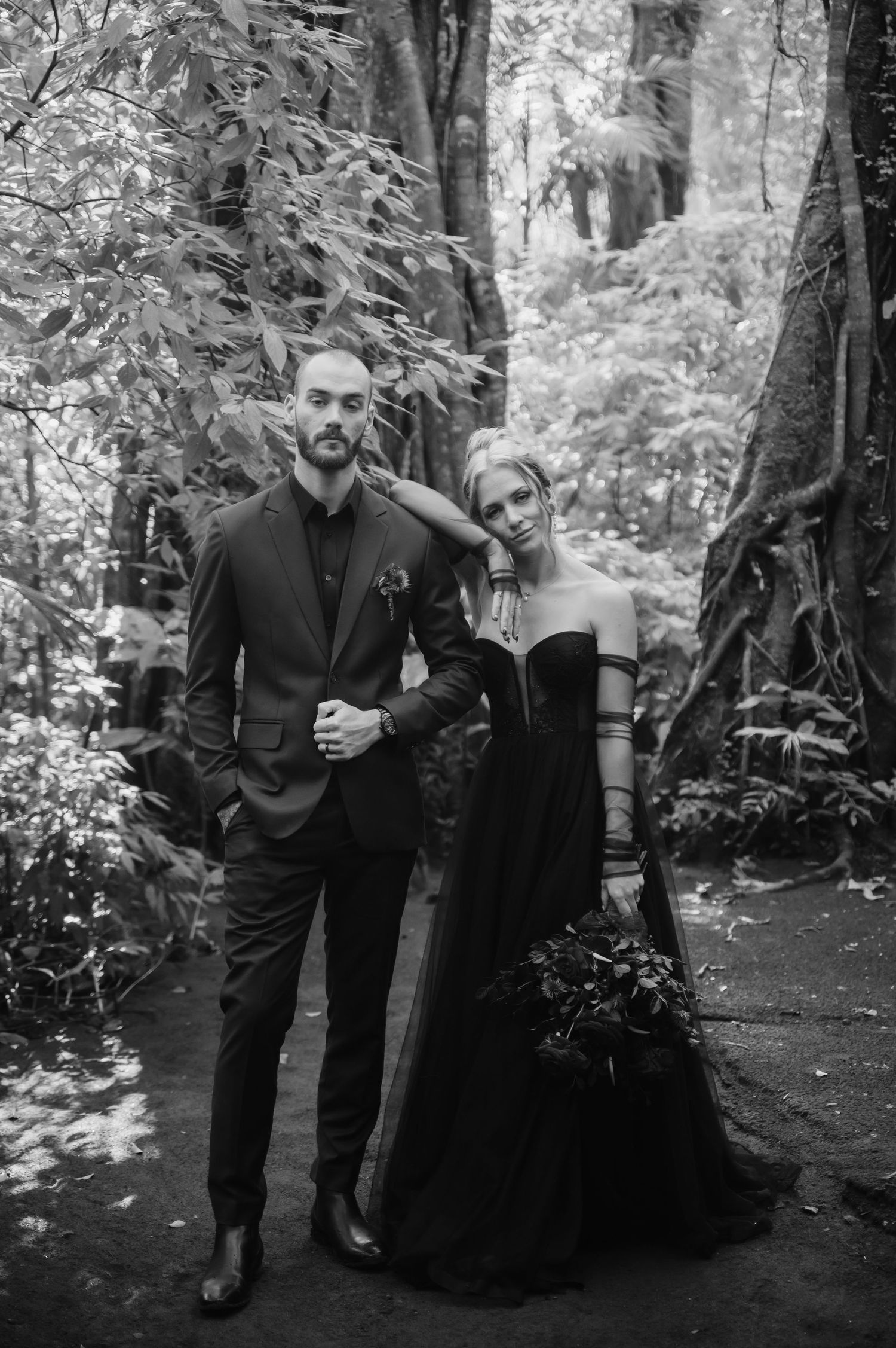 Two people in black formal attire stand together in a forest setting.