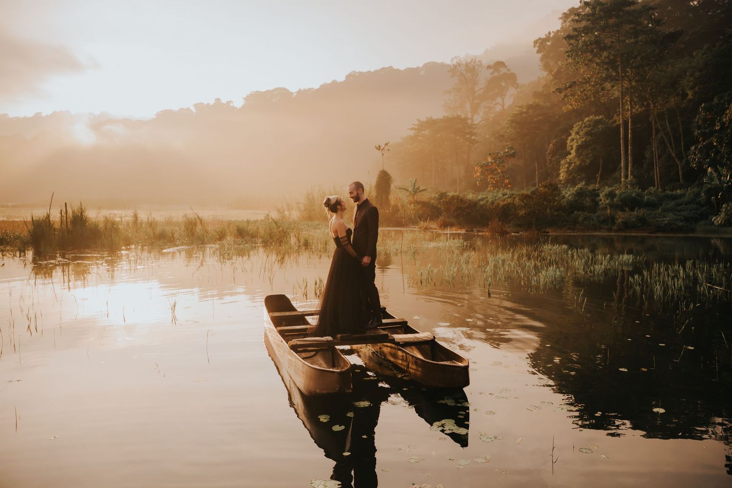 Wide landscape shot of a tranquil lake with boats and figures reflected in the foggy morning light.