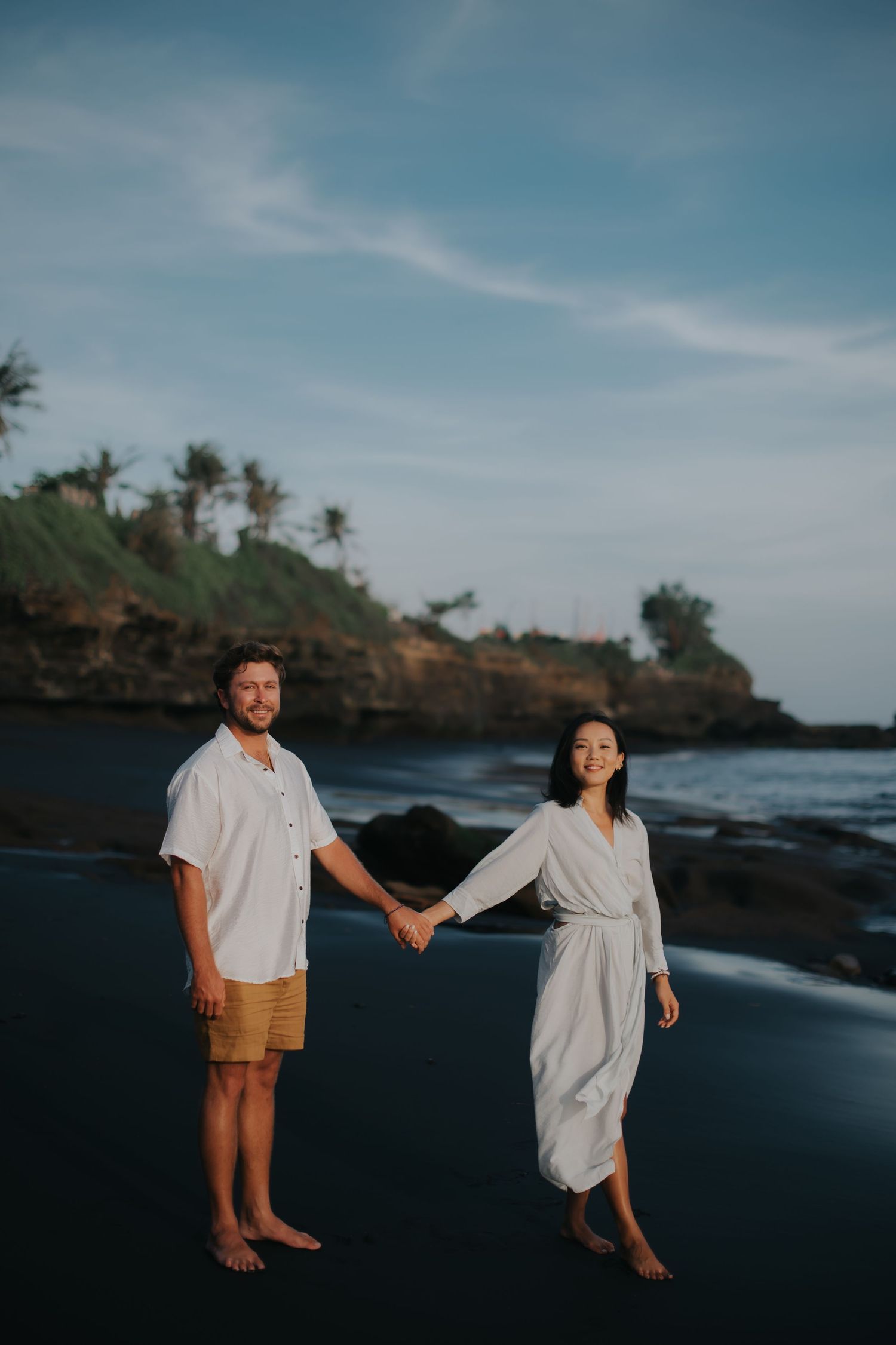 A couple holds hands on a black sand beach with palm trees and rocky cliffs in the background at sunset.