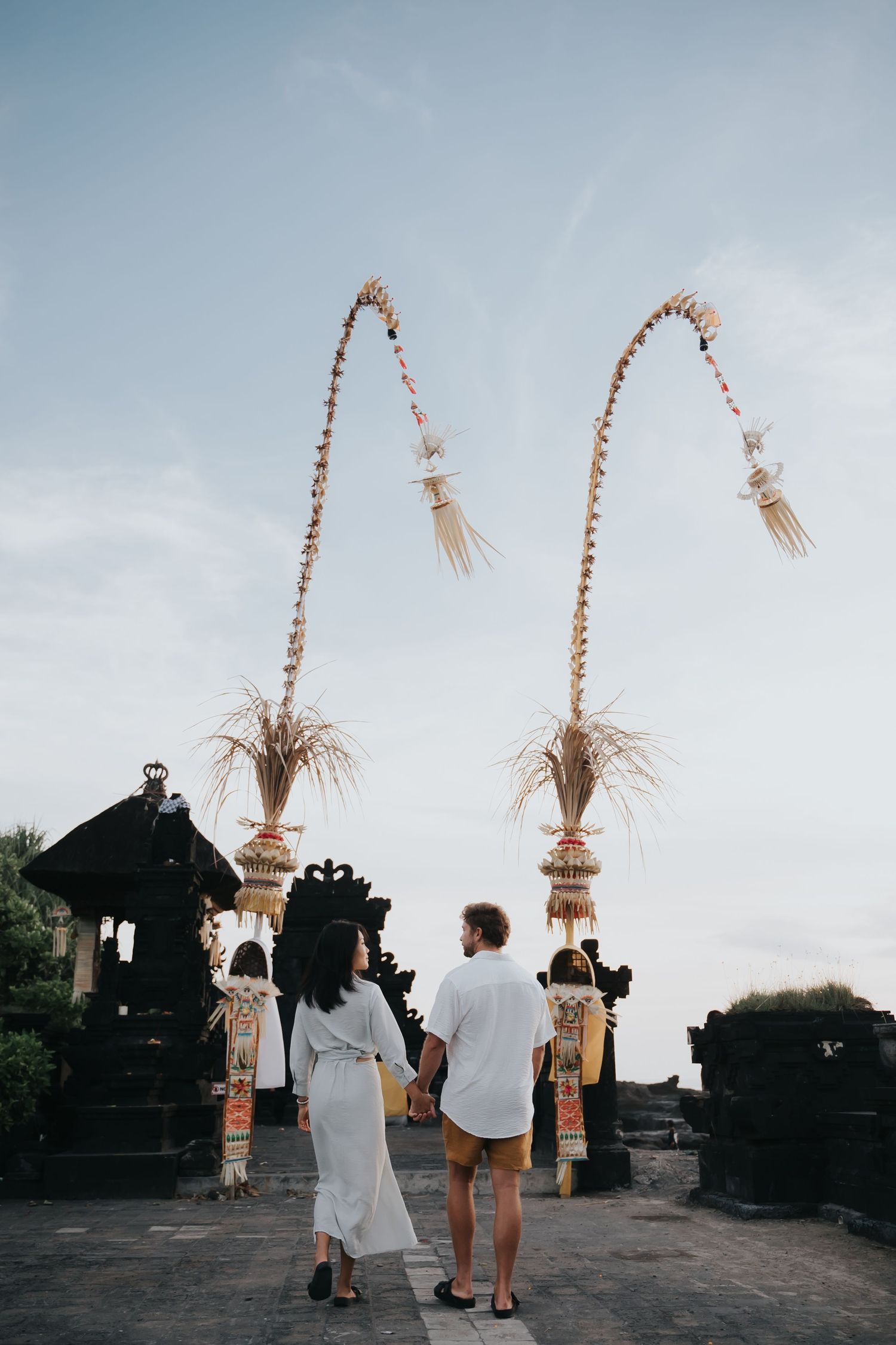Traditional Balinese temple decorations with tall penjor poles against a cloudy sky at sunset.