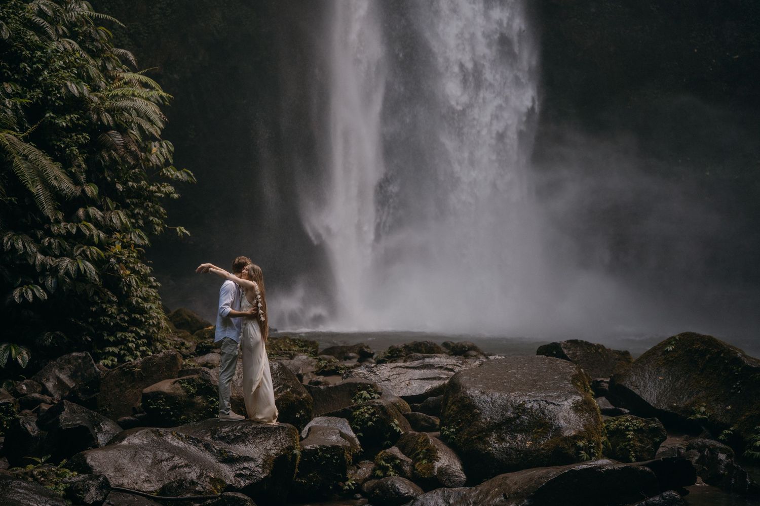 A person stands near a majestic waterfall in a lush forest setting, creating a serene nature scene.