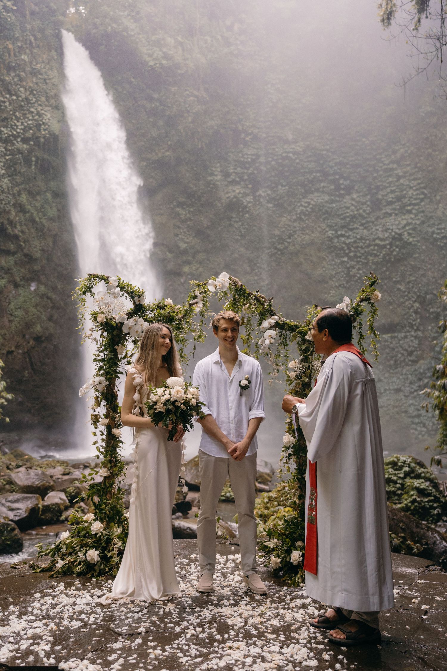 A romantic wedding ceremony takes place beneath a floral arch in front of a majestic waterfall in a misty forest setting.