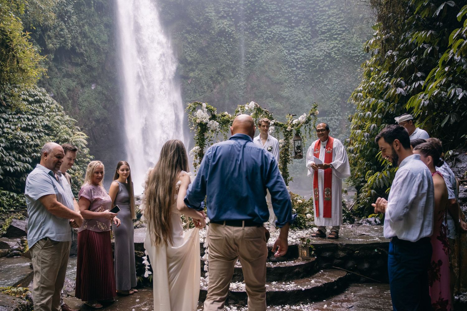 A group of people enjoying a wedding reception celebration outdoors near a scenic waterfall in a lush forest setting.