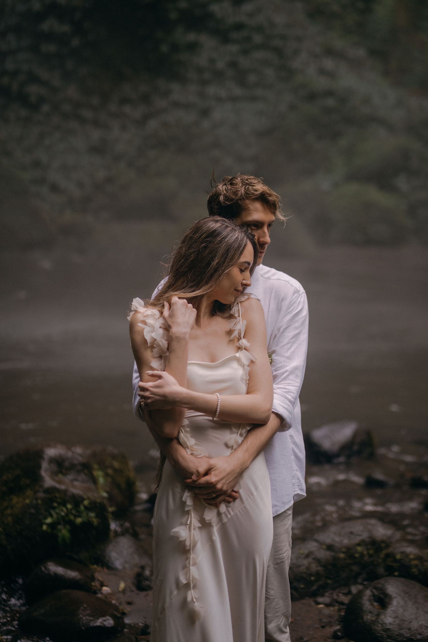 A romantic couple embraces on a misty rocky shore, the bride wearing a flowing white dress and groom in white shirt.