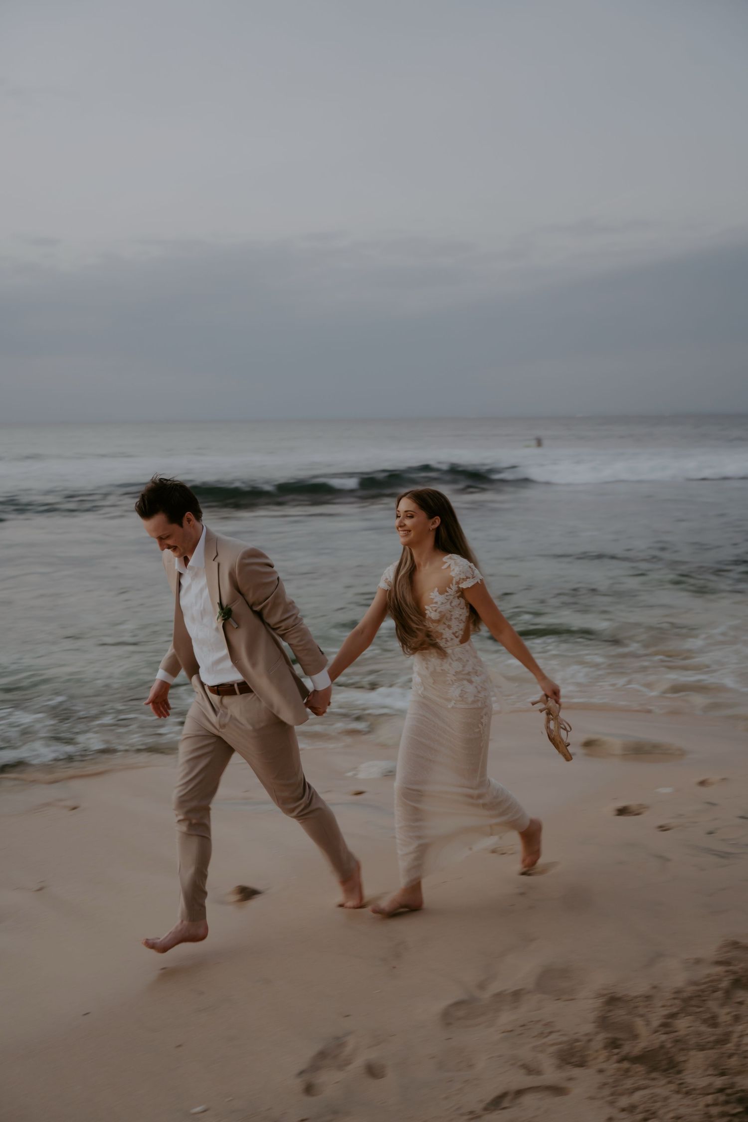 A couple in beige attire playfully runs and dances together on a misty beach at dusk.