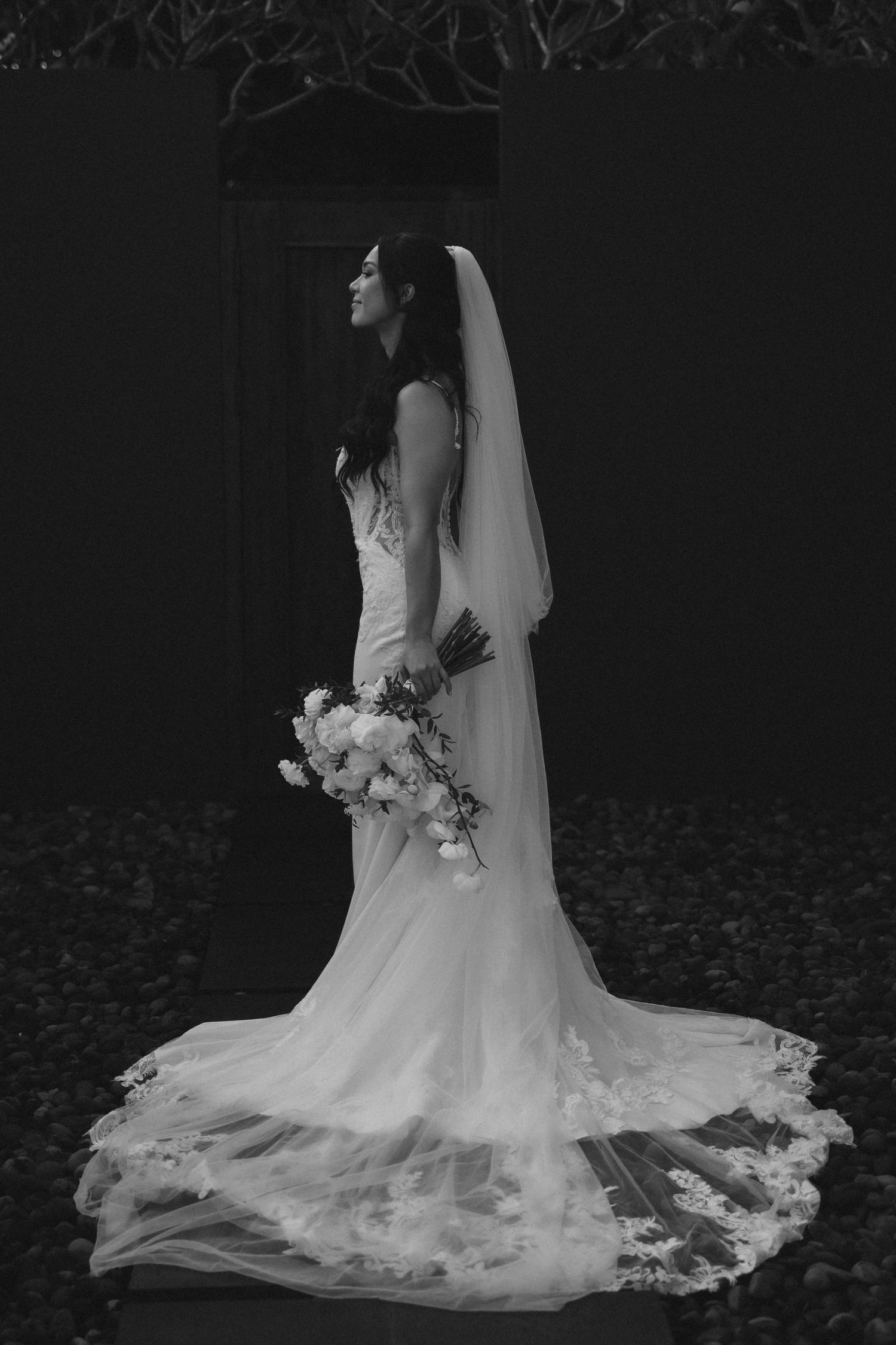 A bride in a flowing white wedding dress with a long train and veil poses elegantly against a dark background.