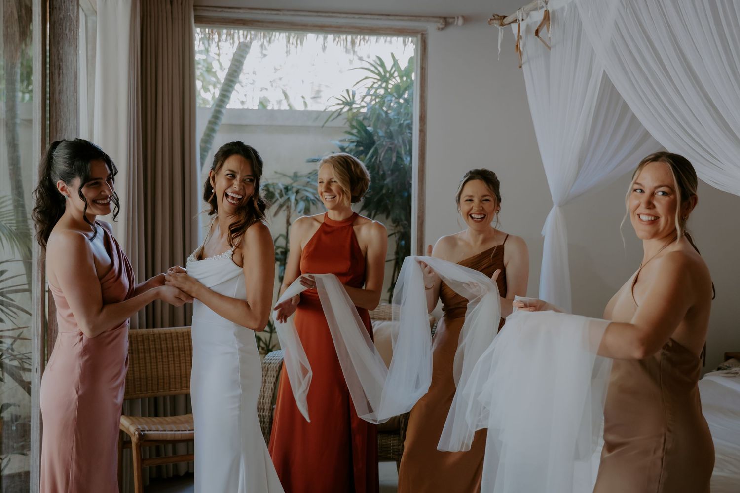 A group of women in formal dresses gather in a sunlit room while helping with wedding preparations.