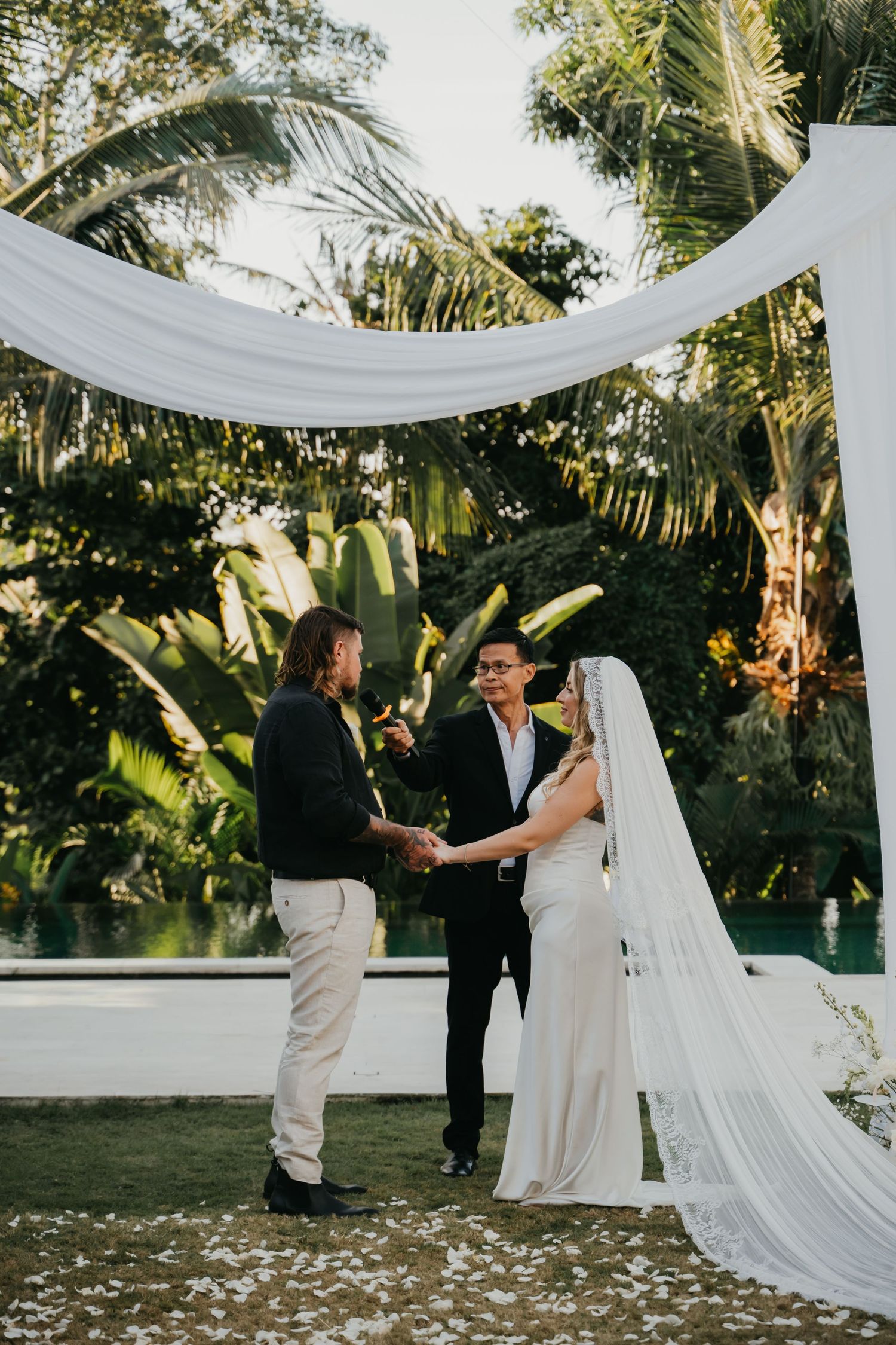 Couple exchanges vows under white draped arch surrounded by tropical greenery at outdoor wedding.