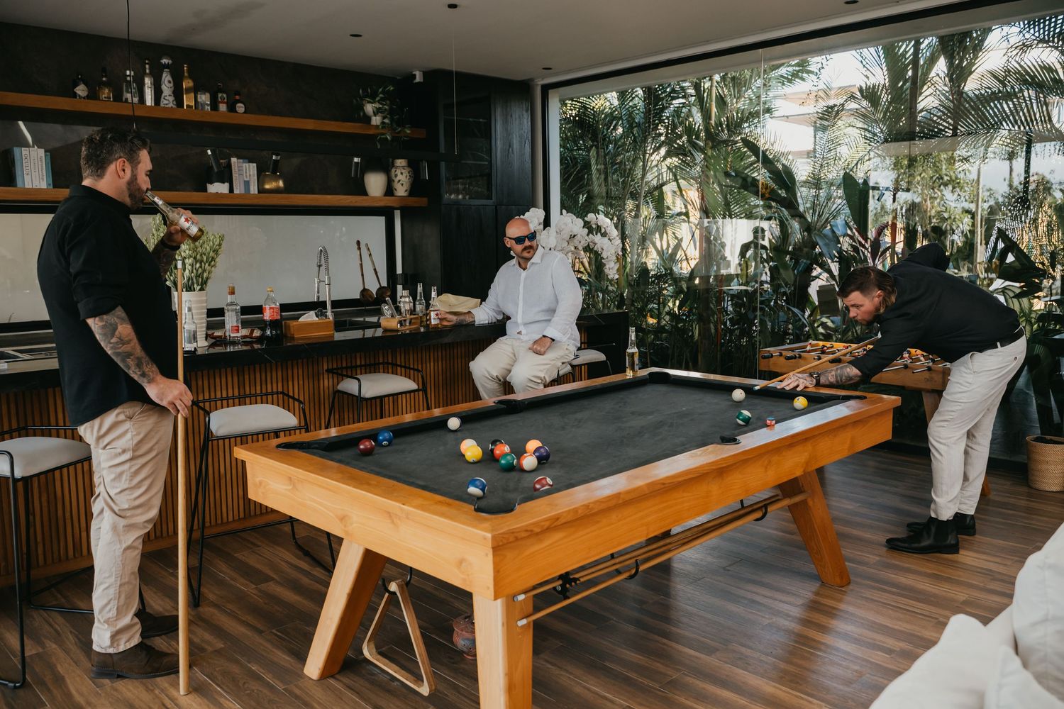 Men playing pool in a modern indoor-outdoor entertainment area with lush tropical greenery visible through large windows.