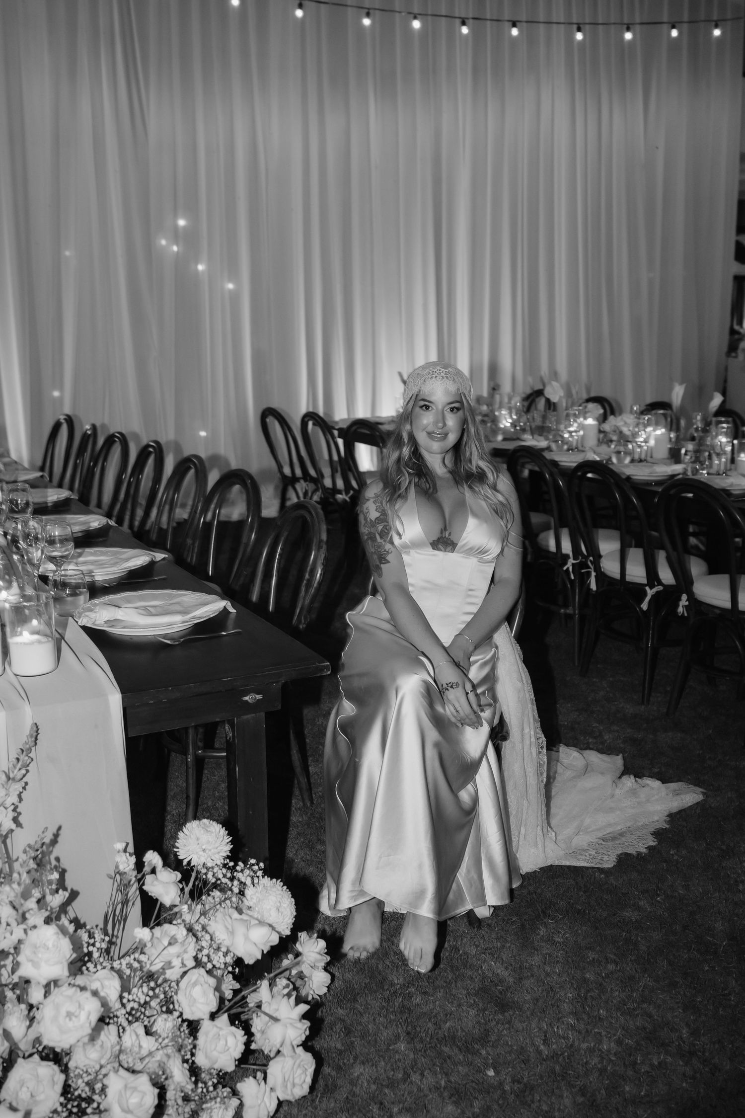 Bride in satin dress sits beside floral arrangements at wedding reception with string lights and white draping.