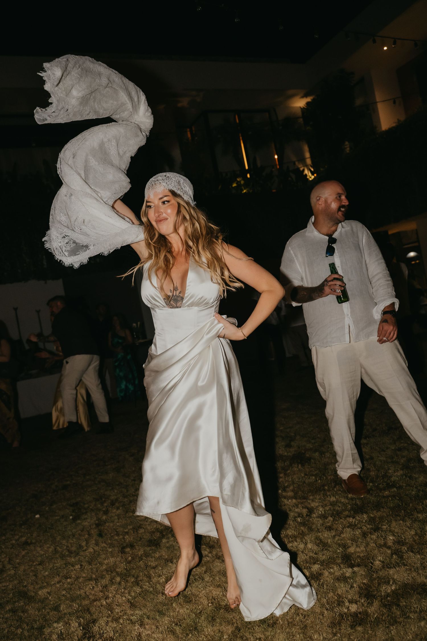 Barefoot bride in satin dress waves veil while dancing at lively outdoor tropical wedding reception.